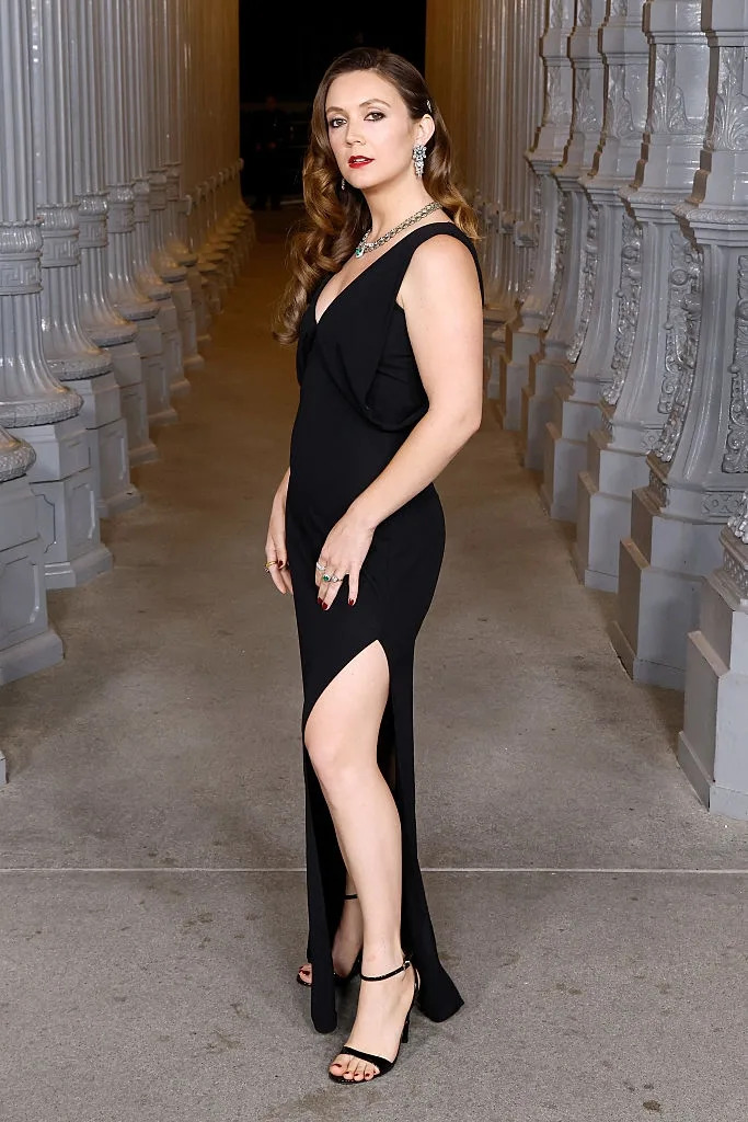 Person in a stylish, sleeveless gown with a thigh-high slit, wearing earrings and a necklace, standing between columns in an elegant setting