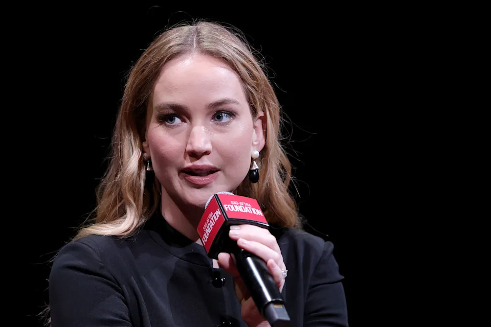 Jennifer Lawrence with long hair speaks into a microphone during a public event, wearing a sleek outfit and pearl earrings