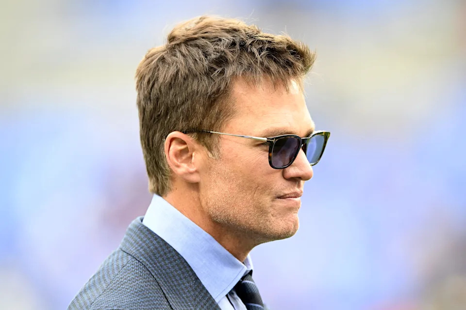 Tom Brady in a suit with sunglasses stands outdoors, facing sideways, with a neutral expression
