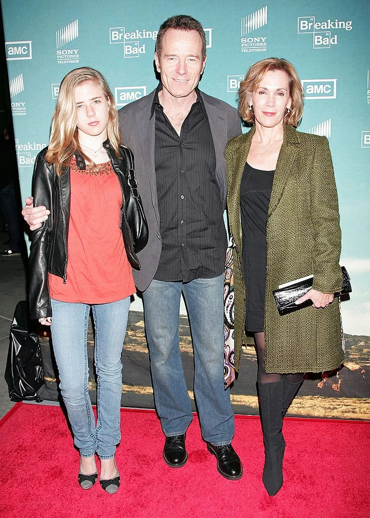 Three people on a red carpet at a "Breaking Bad" event. The middle person is wearing a casual suit, a woman on the right in a coat and dress