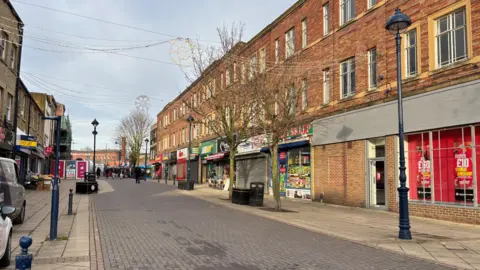 Gemma Dillon/BBC Image shows a pedestrian street.
It is empty and shows several closed or boarded up shops.
There are lamp posts and trees down either side of the street. 
