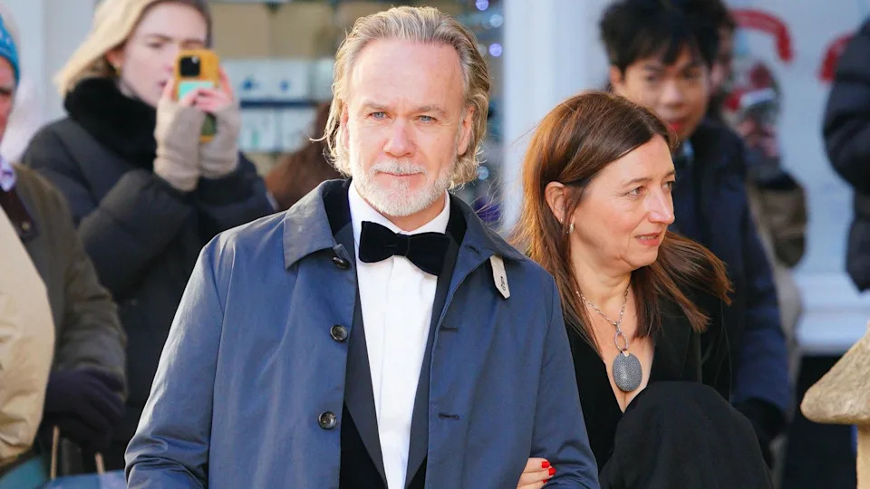A man in a tuxedo and wearing a blue overcoat outside. His partner, a woman dressed in black, is holding his left arm.