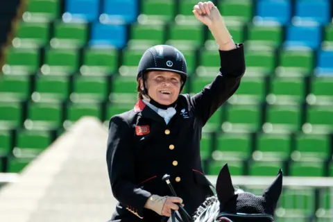 Getty Images Anne Dunham is riding a horse in an arena, wearing a black riding jacket with gold buttons, a white shirt, and a black helmet. The rider’s right arm is raised in a celebratory gesture, holding a riding crop in the left hand. The horse is partially visible, wearing a black ear bonnet. The background shows rows of green and blue stadium seats, indicating a competitive event setting.