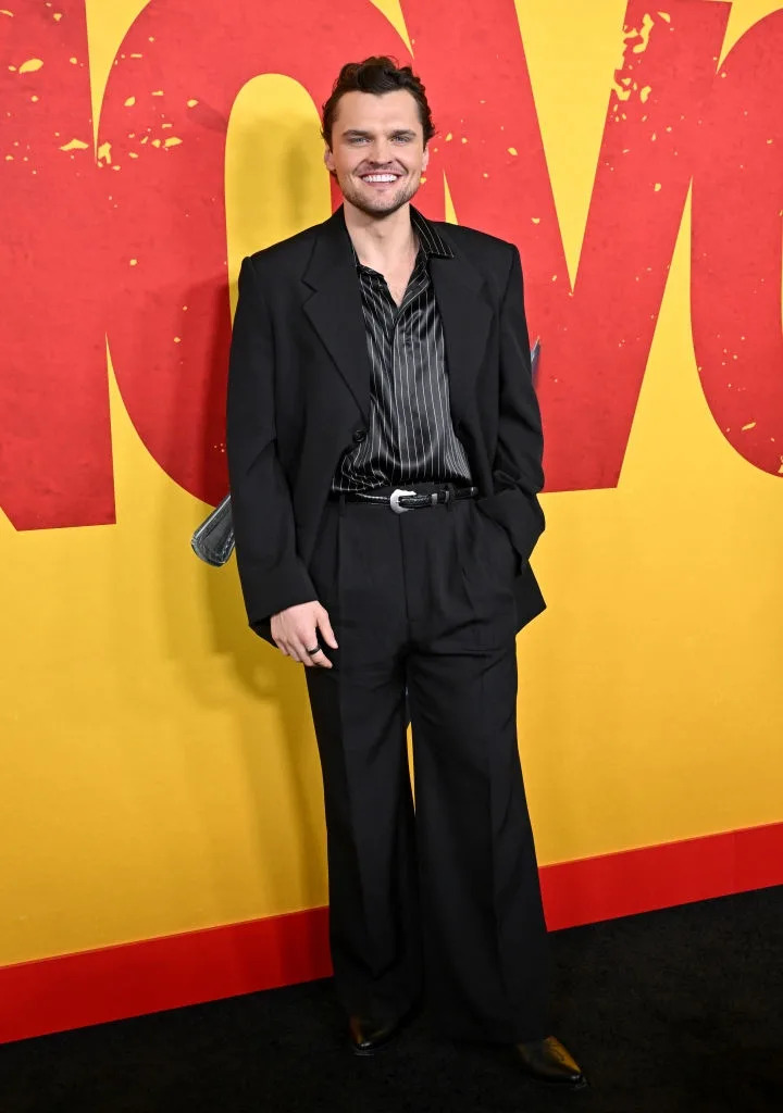Person on a red carpet wearing a black suit with an open-collared striped shirt, smiling