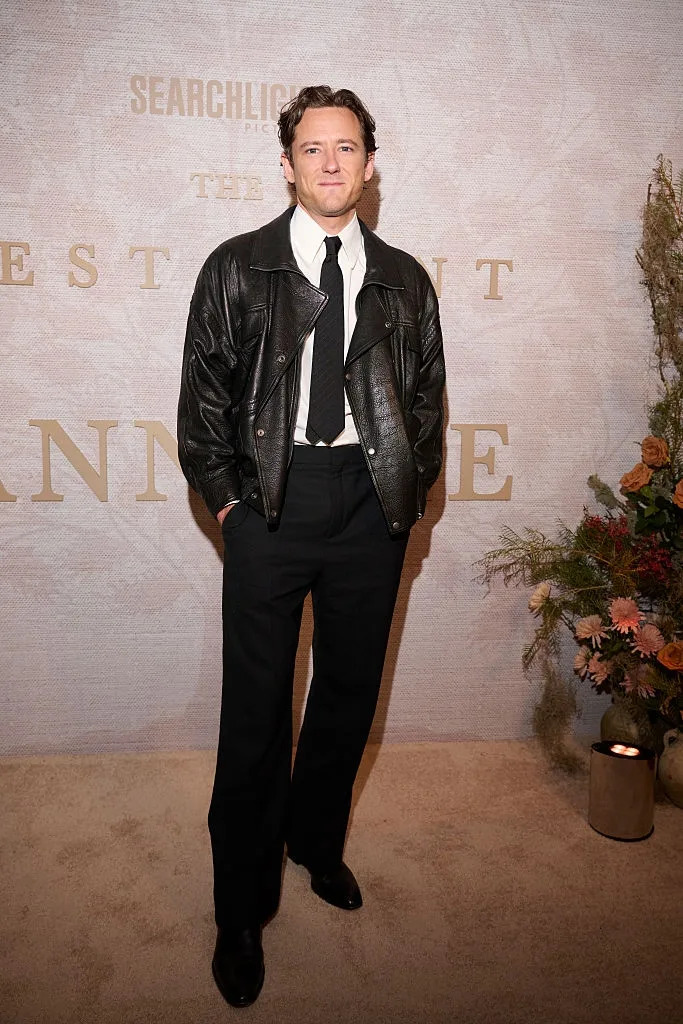 Person in a leather jacket and tie stands on a carpeted area with floral decor and a textured backdrop