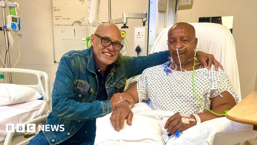Garry Christian, smiling wearing black glasses and a denim jacket, clasps the arm of Lionel Duke, who is also smiling while sitting up in a hospital chair, wearing a patient's gown, with various tubes attached to parts of his body.