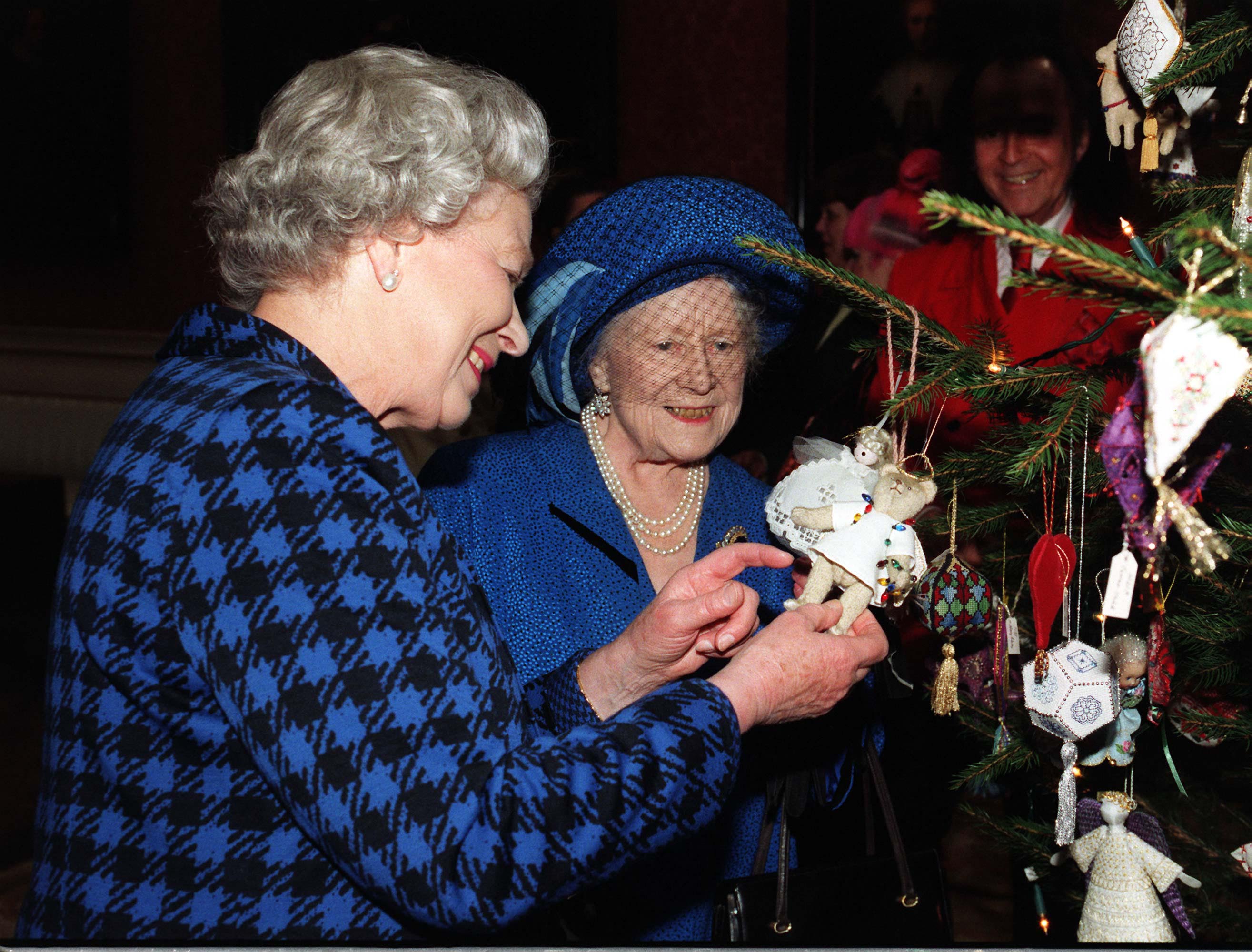 Queen Elizabeth and the Queen Mother looking at ornaments on a Christmas tree, both dressed in blue coats