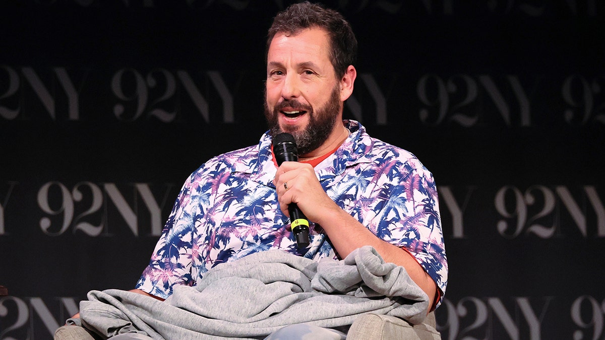 Adam Sandler sits down in a purple and blue patterned shirt with a sweatshirt on his lap, speaking into a microphone on stage