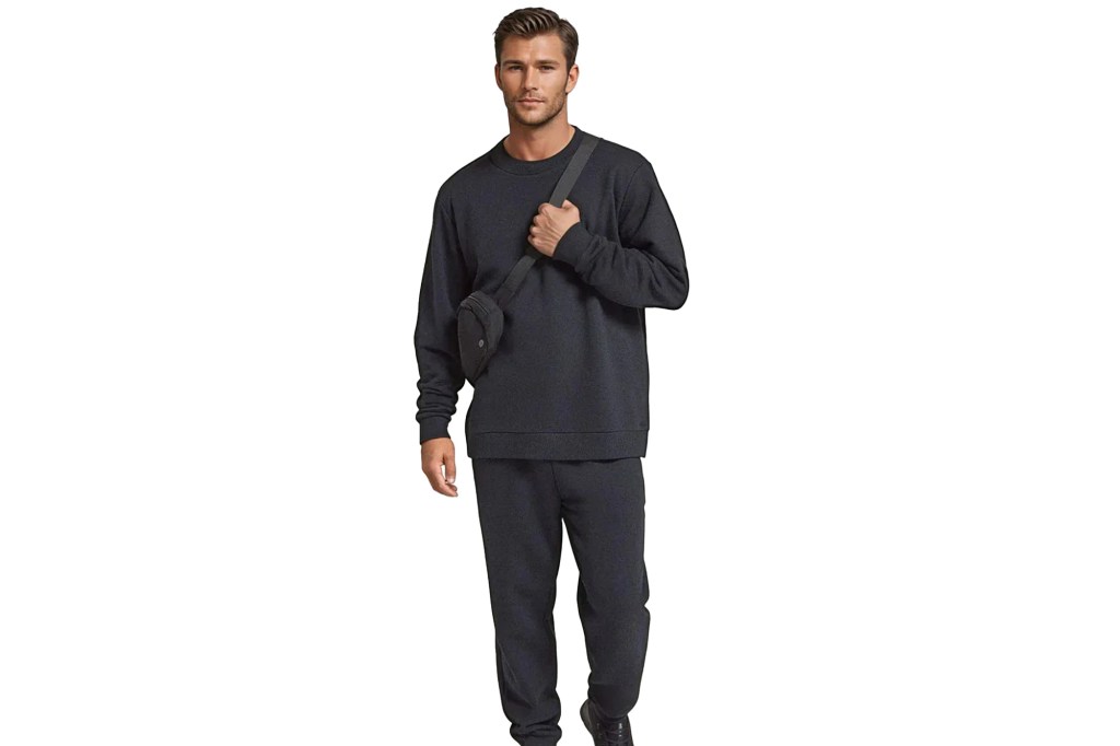 A male model wearing a dark gray sweatshirt and matching sweatpants. He is also wearing a black sling bag across his chest.