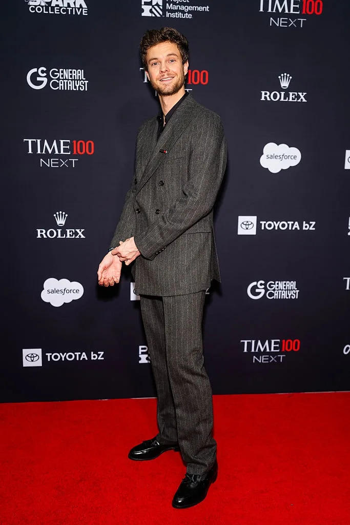 A person in a sharp, pinstriped suit poses on the red carpet at a TIME 100 event, with logos in the background