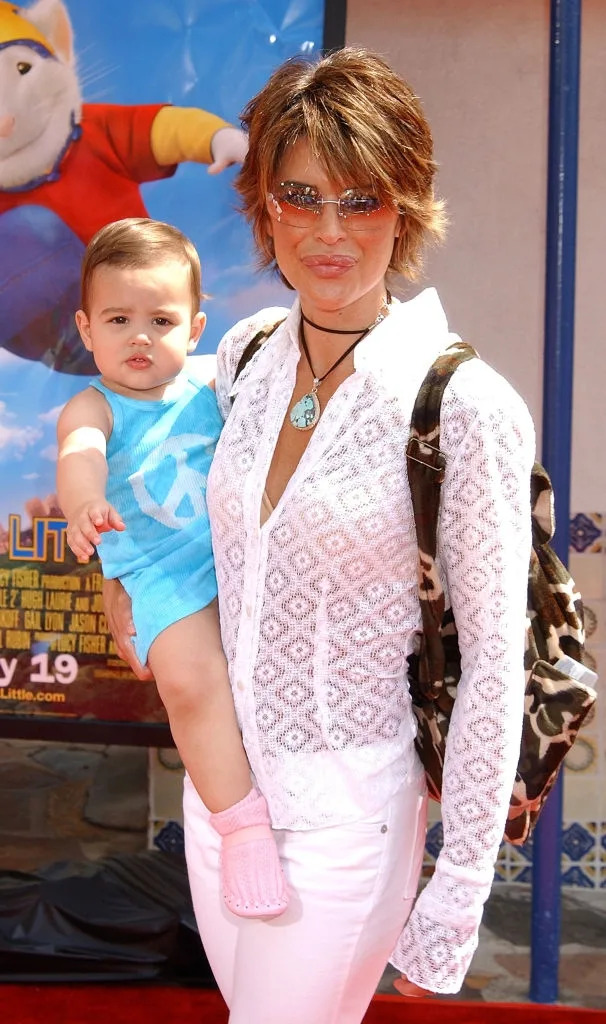 Person holding a baby on a red carpet. The adult wears a sheer patterned top, sunglasses, and a backpack. The baby is in a casual outfit