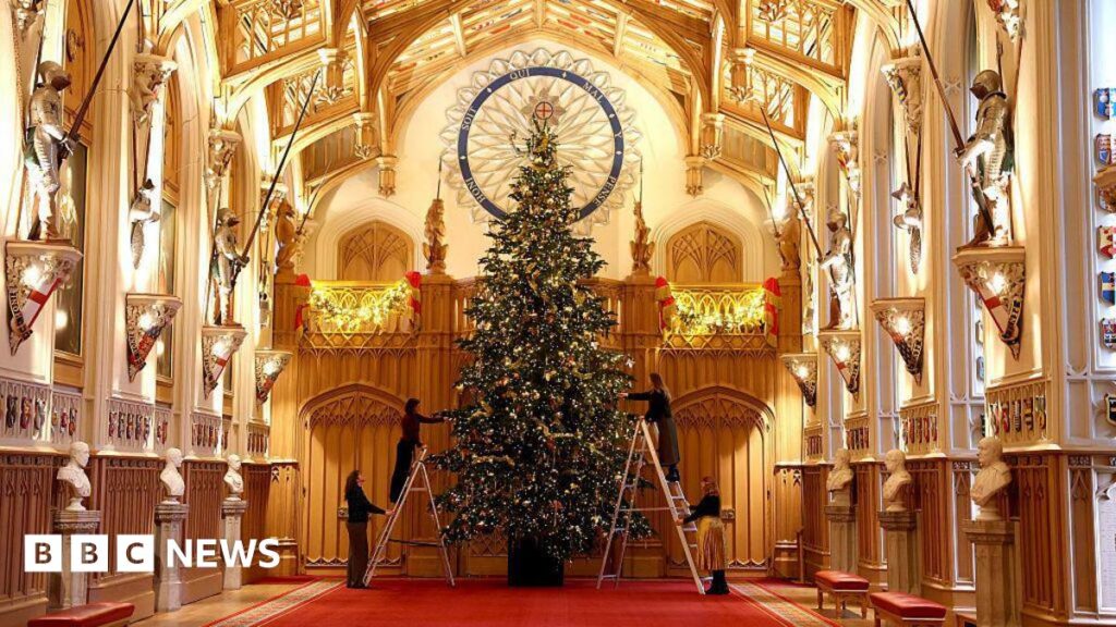 There is a grand corridor with a gold ceiling. A Christmas tree is positioned in front of it, with two people putting up the decorations on the tree on a ladder, with someone holding each ladder.