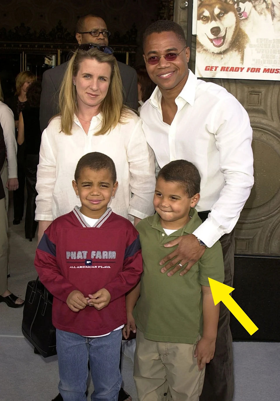 A family poses together at an event. The man in sunglasses wears a casual shirt, and the woman next to him wears a white blouse. Two children stand in front