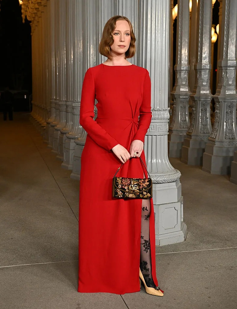 A person in an elegant long gown with floral lace accents on one leg, holding a detailed handbag, stands in front of an outdoor column display