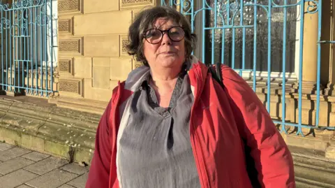 Gemma Dillon/BBC A lady wearing a red jacket and grey top is standing in front of some blue rails. 
She has brown short hair and glasses.