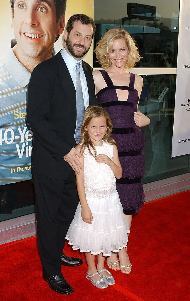 A family poses on the red carpet; the woman wears a sleeveless dress, the man is in a suit, and the child is in a white dress