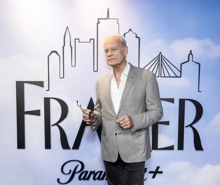Person in a grey suit stands in front of a "Frasier" backdrop with skyline design, holding glasses