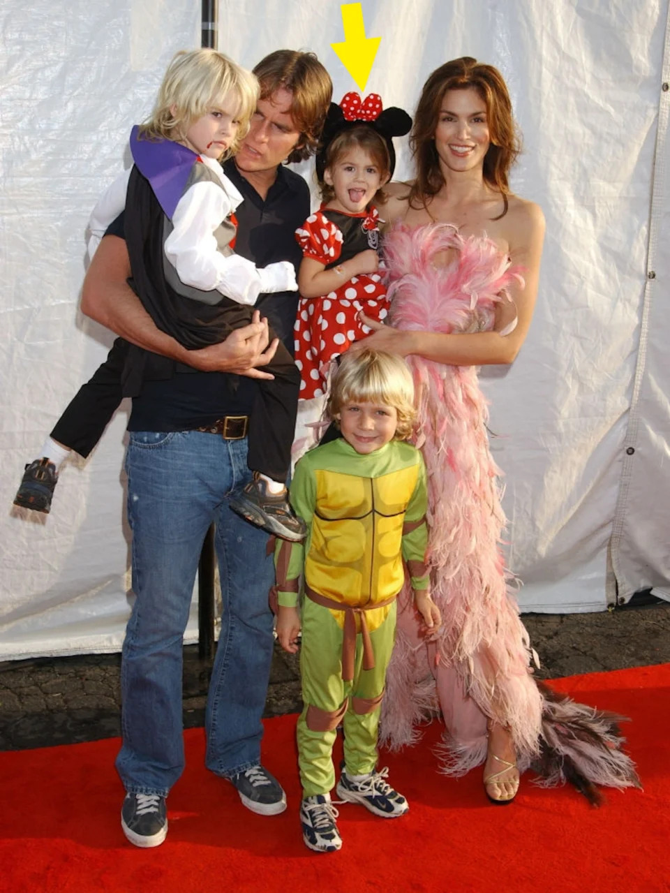 A family poses together on a red carpet. The woman wears a feathered gown, the man is casual, and the children are in costume, including a turtle and a mouse