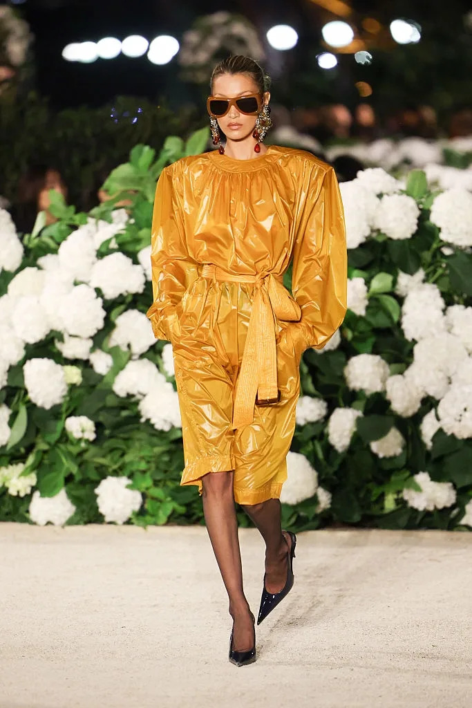 Person on runway in chic oversized shiny outfit, statement earrings, and sunglasses, surrounded by white flowers and greenery