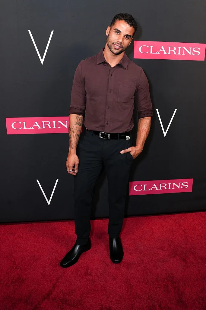 Person posing on red carpet in a casual outfit with a short-sleeved shirt, black pants, and shoes, standing in front of a branded backdrop