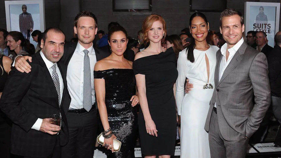 Meghan Markle pictured with her Suits co-stars at an event in New York in 2012. From left to right: Rick Hoffman, Patrick J. Adams, Meghan Markle, Sarah Rafferty, Gina Torres and Gabriel Macht. Ms Markle wears a black sequined off-the-shoulder dress and carries a gold clutch bag. 