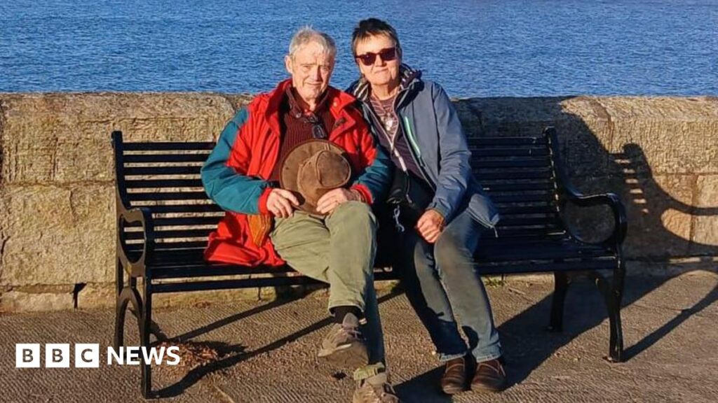The North Yorkshire names on the King’s New Year Honours list Ruth and her husband sit on a bench by a low stone wall, against a backdrop of the sea. They both wear waterproof coats. Ruth has short dark hair and wears sunglasses.