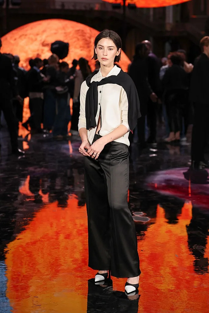 Person in stylish attire with a shawl and wide-leg pants stands on a reflective surface at an event with a vibrant backdrop