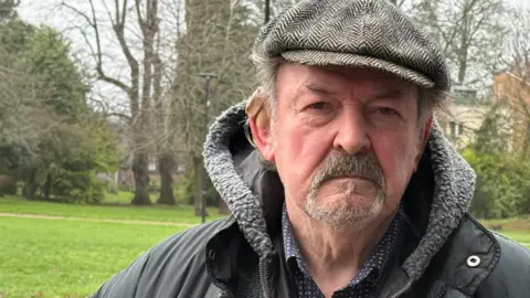 Danny Fullbrook/BBC An older man wearing a dark jacket with a fleece-lined hood and a flat cap is standing outdoors.
