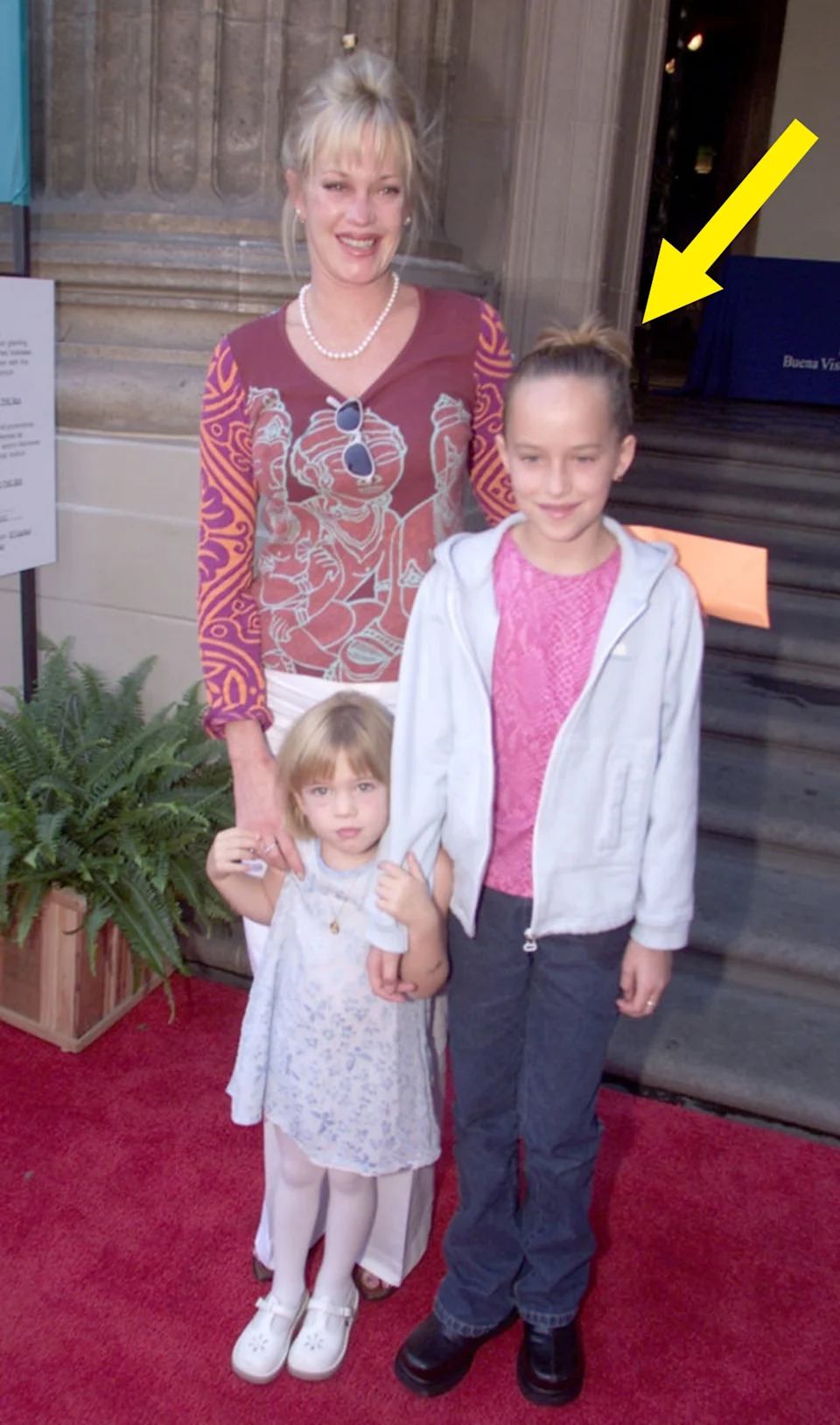 A woman in a patterned top and pearls poses on a red carpet with two young children, one in a light dress and the other in casual clothes