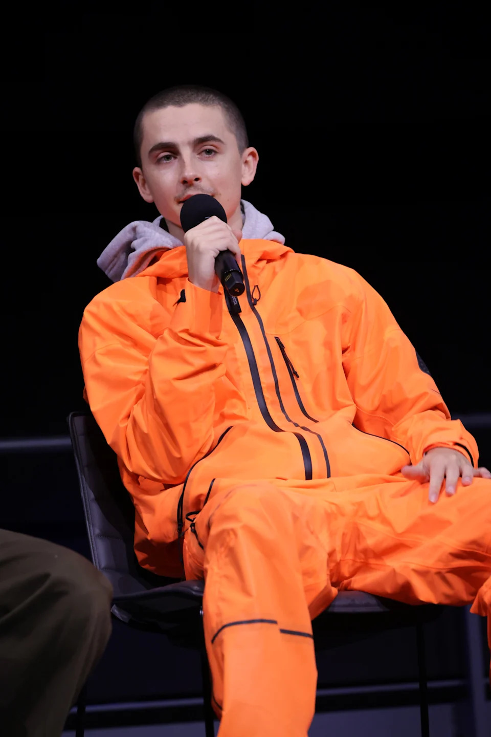 Timothee Chalamet in a jumpsuit with a hood speaks into a microphone while seated on stage at a public or media event