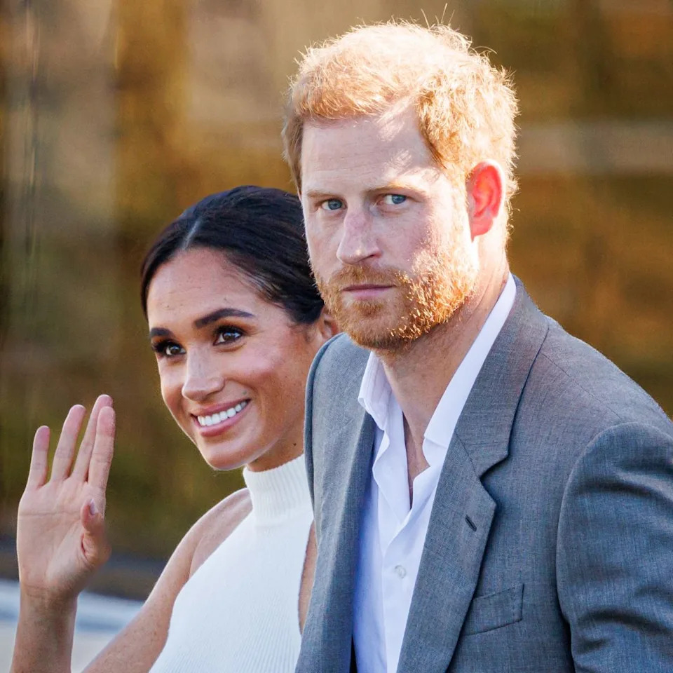  Meghan Markle wears a white halter top and waves while Prince Harry, who has red hair, wears a gray suit jacket and white shirt. 