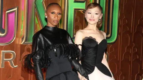 Getty Images Cynthia Erivo in a black top with leather or leather-look sleeves and feathers and Ariana Grande in a corset-style strapless dress which is black at the top with a taffeta white skirt. They are at the New York premiere of Wicked: For Good.
