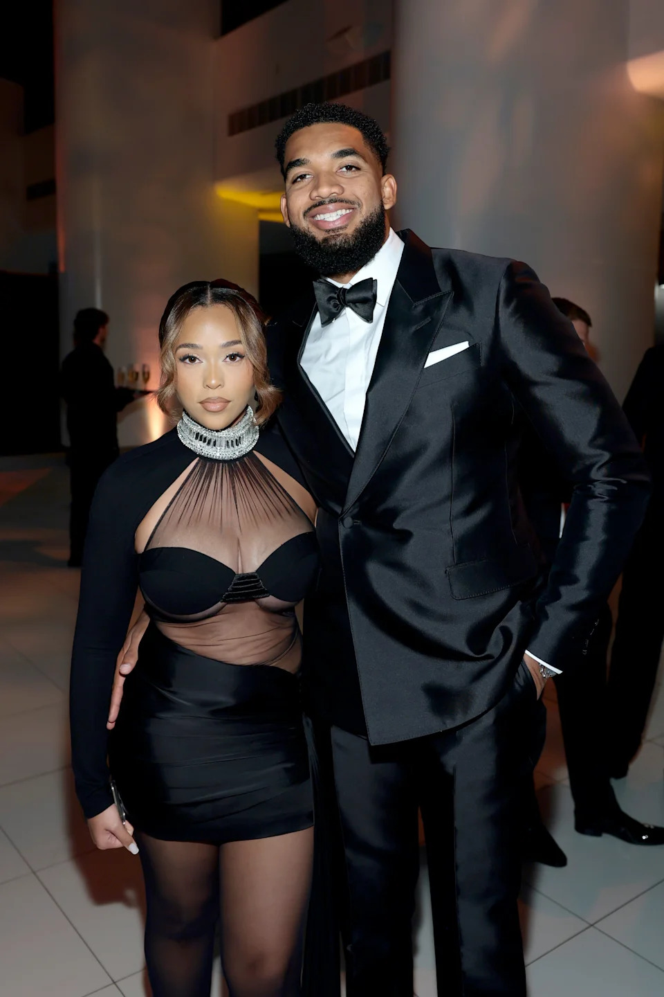 Karl Anthony Towns in a tuxedo and Jordyn Woods in a sleek dress pose together at an event, showcasing elegant evening wear