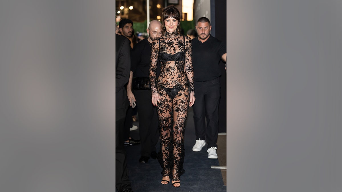 Dakota Johnson poses at the Kering Foundation Caring for Women Dinner wearing a floor-length sheer black lace gown with a high neckline and visible underlayers.