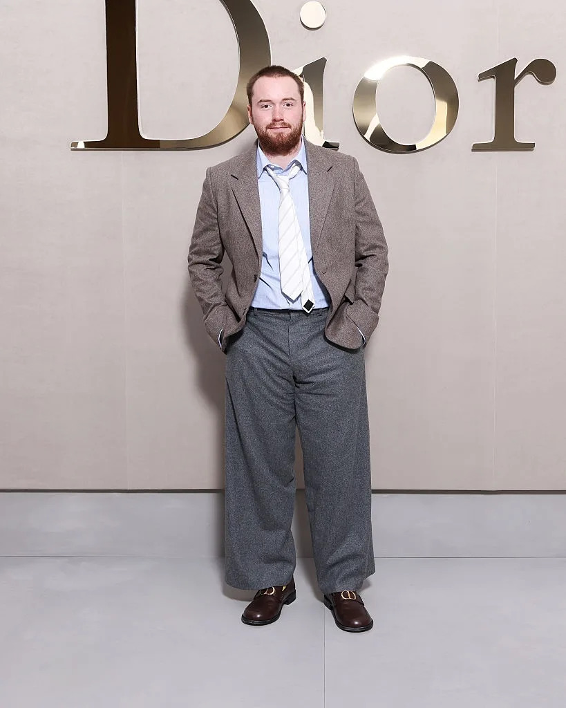 Person posing in front of a Dior logo in a vintage-style outfit: oversized suit jacket, wide trousers, light shirt, and tie