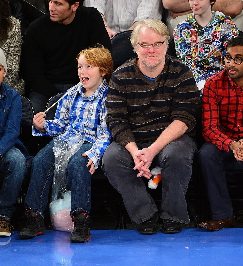 Two people sitting courtside at a basketball game, one in a striped sweater and glasses, the other in a plaid shirt eating cotton candy