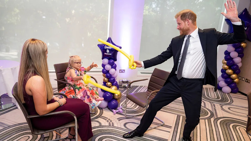 At the WellChild awards in London in September 2025, a smiling Prince Harry wears a dark suit and tie while holding a yellow balloon sword and playing with nine-year-old Gwen Foster, who received the Inspirational Child 7-11 award. Gwen wears a multi-coloured dress and glasses and is also holding a yellow balloon sword. 