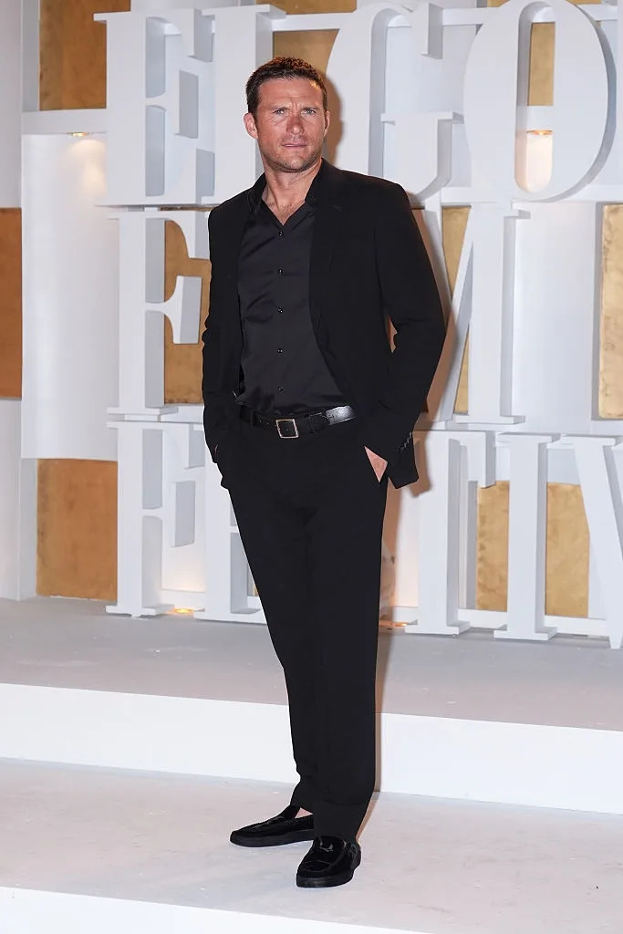 Man in a black suit with no tie on a red carpet, standing in front of large 3D letters