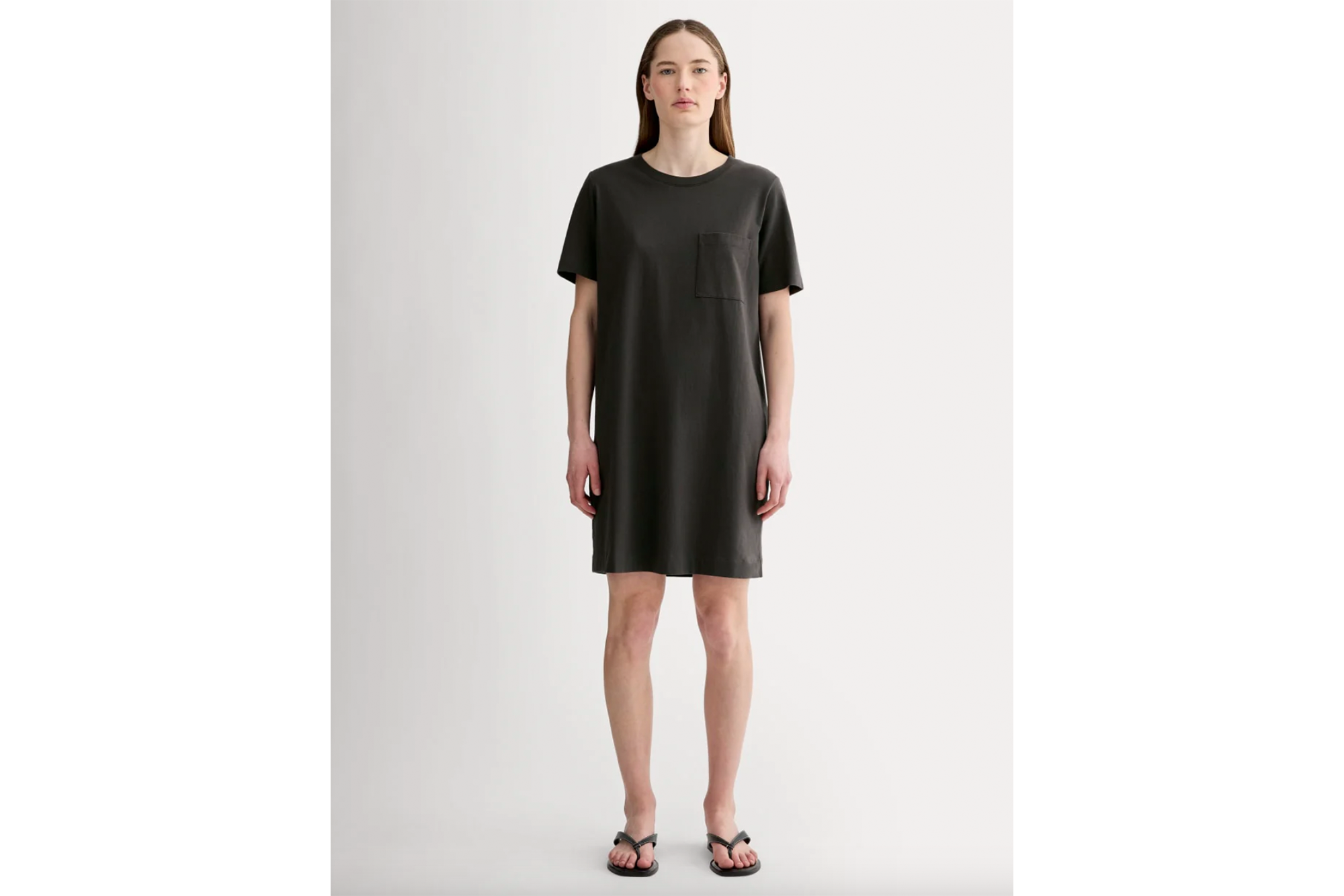 A woman wearing a dark grey t-shirt dress with a left chest pocket and black flip-flops.