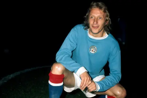 Getty Images Denis Law is kneeling on one knee on a pitch, wearing a long-sleeved light blue Manchester City jersey with a circular badge on the chest, white shorts, and red socks with white tops. The background is dark, suggesting the photo was taken during night-time or under stadium lighting. Law's hands are resting on the bent knee, and the overall setting indicates a posed shot before or after a match.