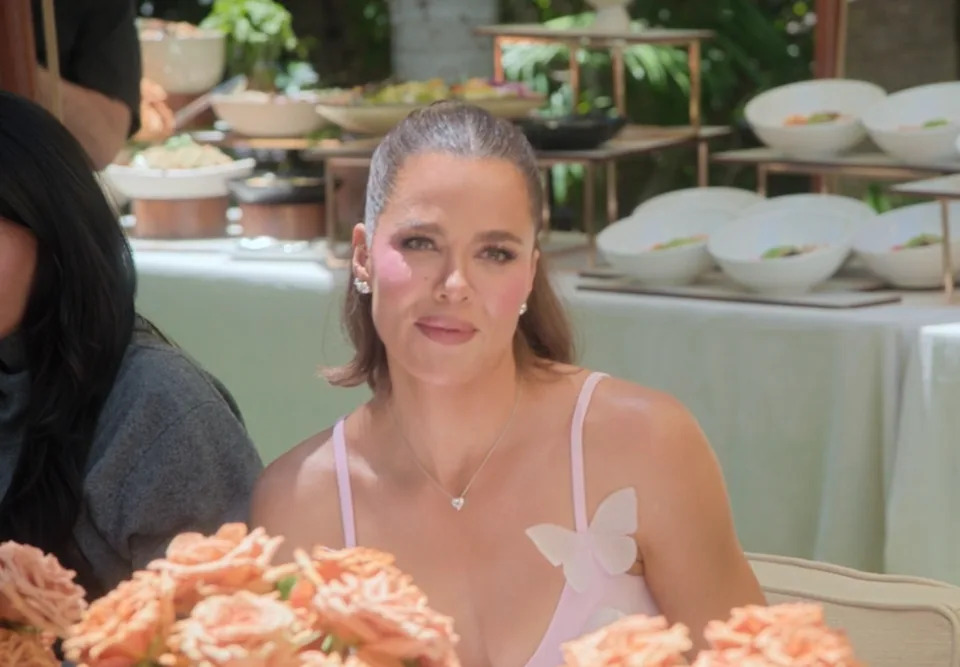 Khloé seated at an elegant event with floral decor, pulling a face