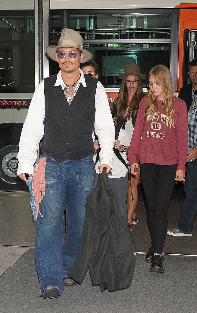 A group of people walk at an airport. The person in front wears a hat, sunglasses, vest, and scarf. Others wear casual outfits and hats
