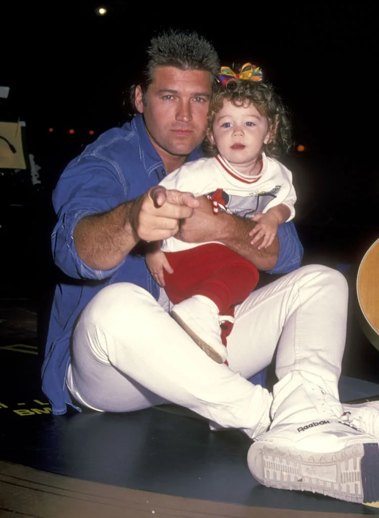 A person in a casual outfit poses with a child on their lap, pointing towards the camera. The child is wearing a festive bow and casual clothing