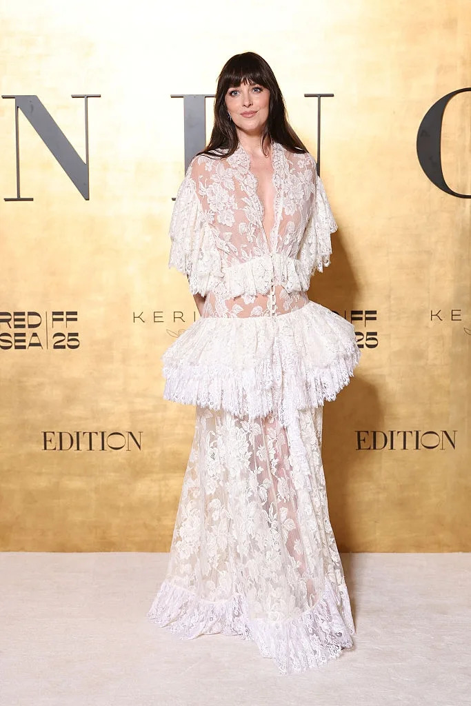 Person in a sheer, floral lace dress with layered ruffles poses on a red carpet against a gold backdrop with text