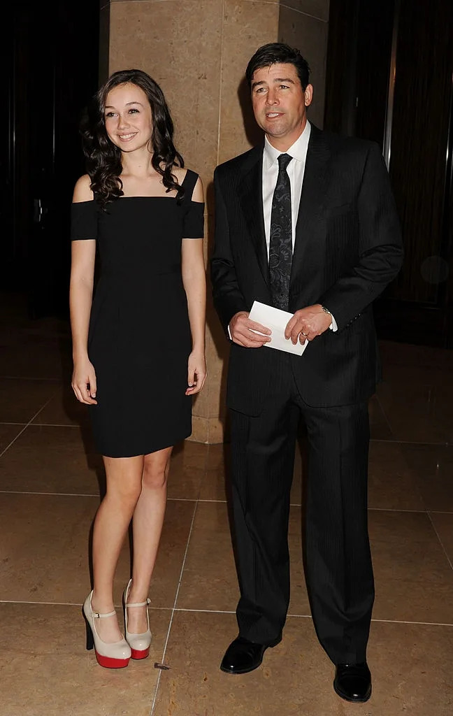 Two people pose at an event. The person on the left wears a black off-shoulder dress, while the person on the right wears a dark suit with a tie