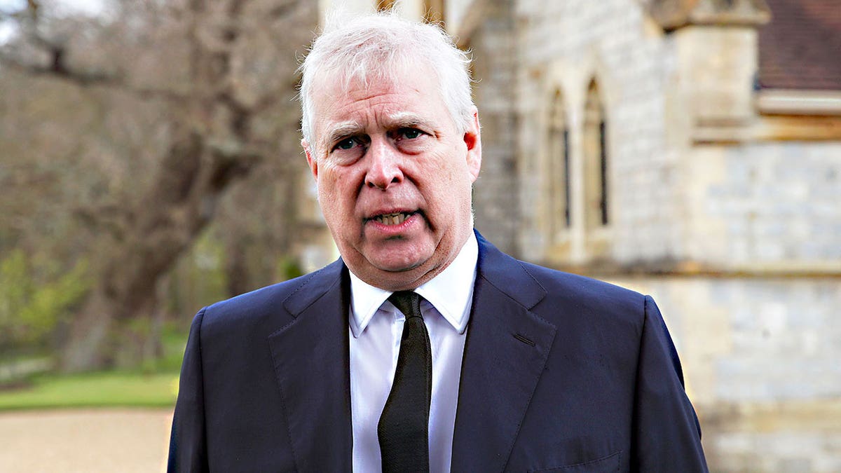 Prince Andrew looking distressed in a dark suit and tie in front of a church.