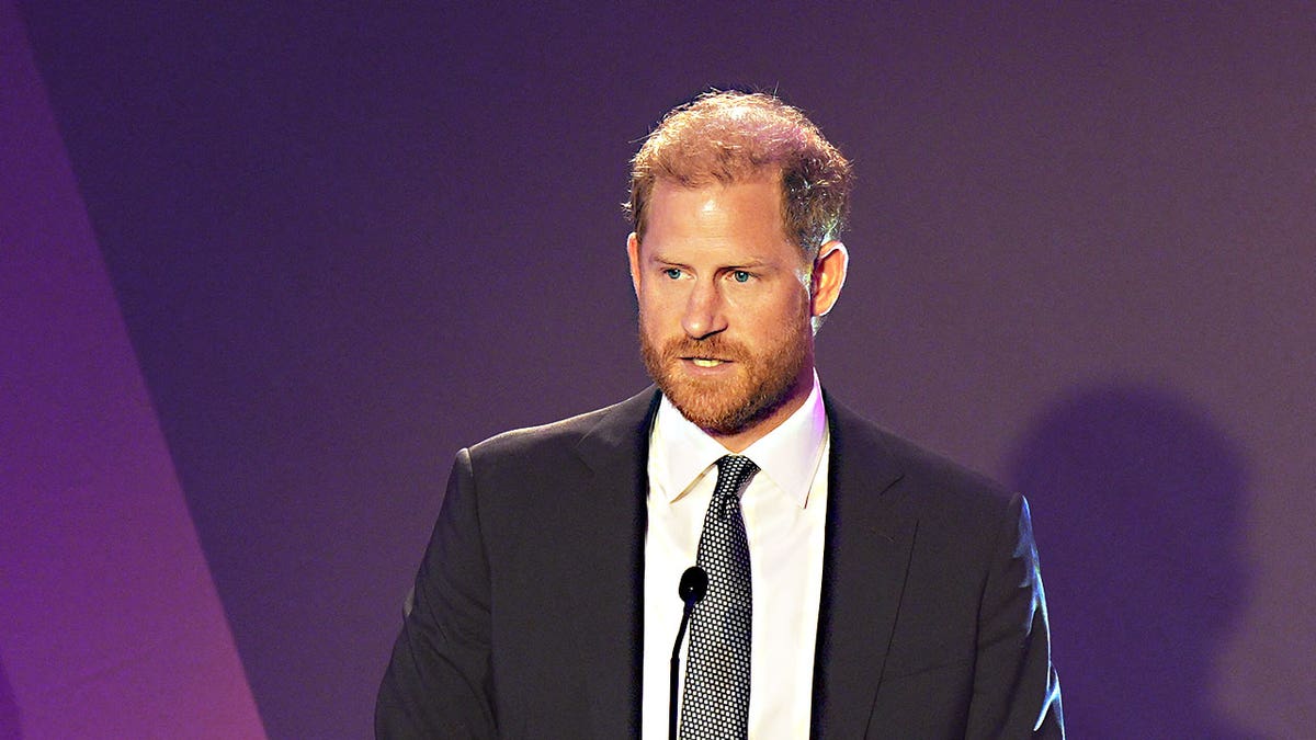 Prince Harry in a suit speaking at a podium looking serious.
