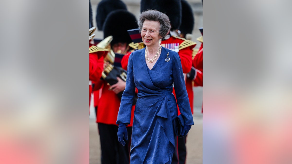 Princess Anne wearing a blue dress walking in front of British guards and smiling.