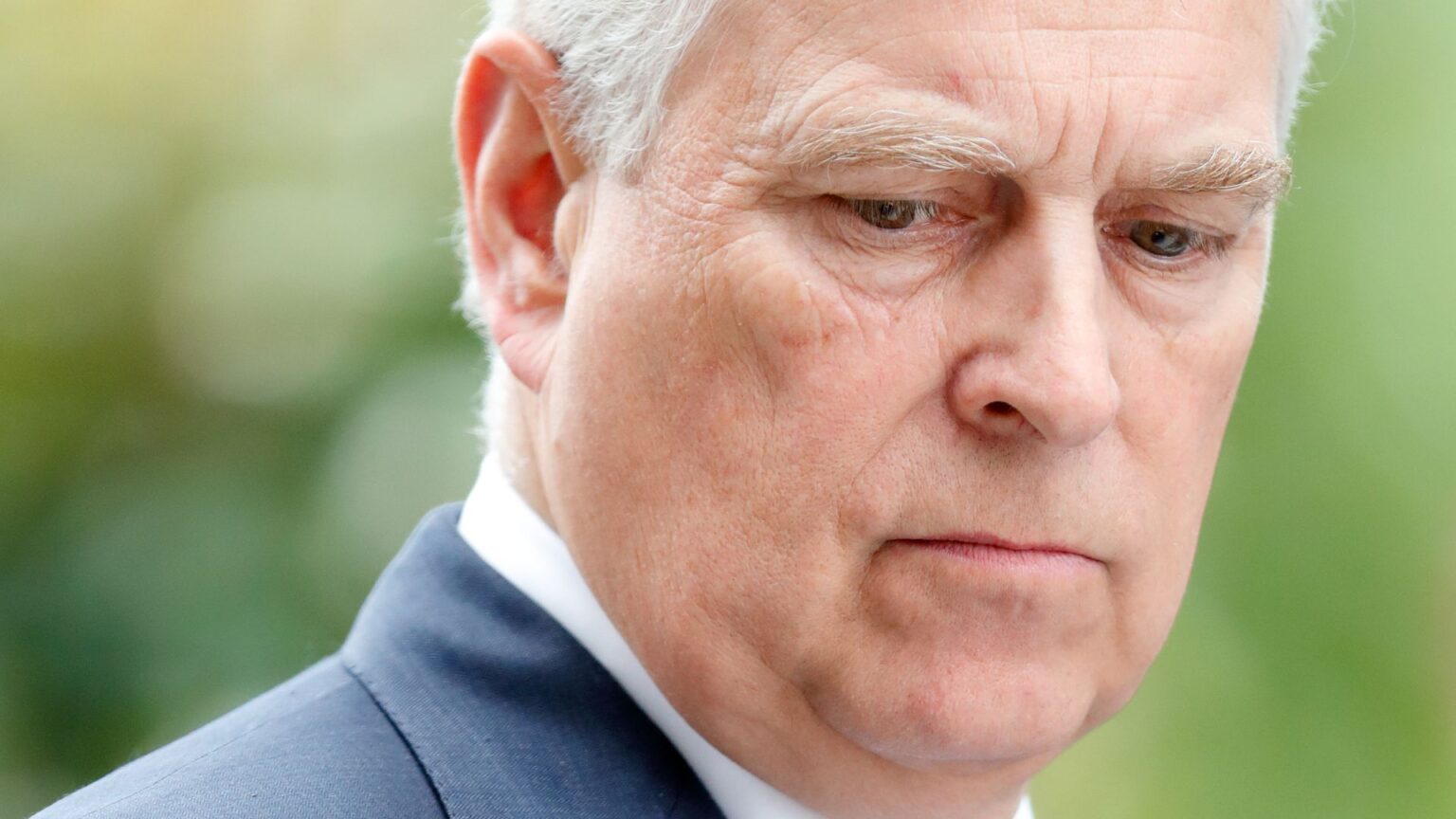 Royal Author Claims Ex-Prince Andrew Is Delaying Downsizing A closeup picture of Prince Andrew looking upset and wearing a blue suit with a white dress shirt and red tie