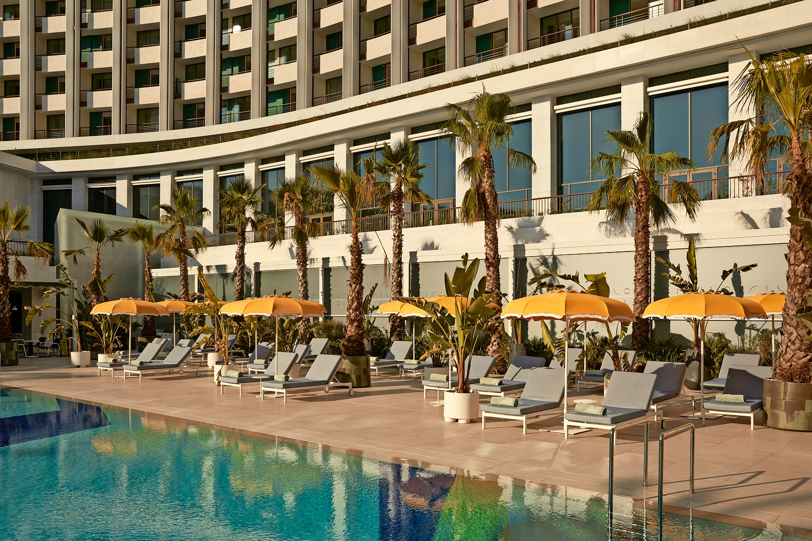 Outdoor pool area with lounge chairs, yellow umbrellas, and palm trees at Conrad Athens, The Ilisian.